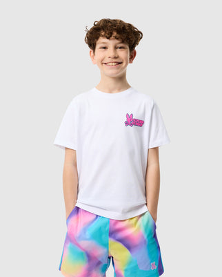 A young boy with curly hair smiling, wearing a white Psycho Bunny Pima cotton T-shirt with a pink bunny logo and colorful tie-dye shorts, standing against a light grey background.