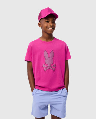 A smiling young boy wearing a bright pink KIDS RIVIERA GRAPHIC TEE - B0U639C200 by Psycho Bunny, featuring a large bunny and crossbones graphic, a matching pink cap, and light blue shorts. His hands are in his pockets, and he is standing against a plain white background.