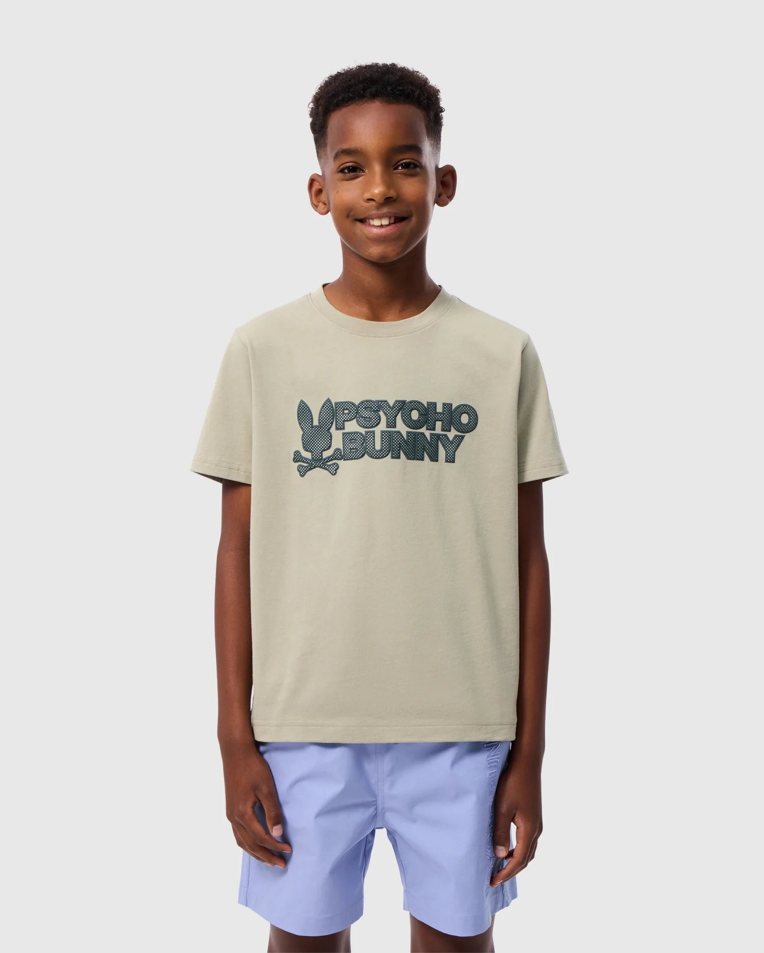 A young boy smiling and standing confidently. He is wearing a light beige Pima cotton t-shirt with the text 