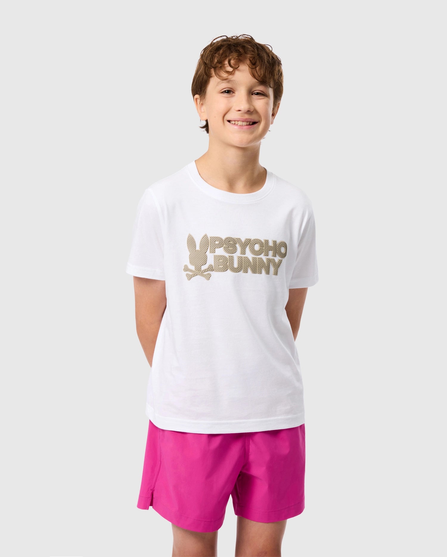 A smiling boy stands against a plain background, wearing the Kids Melvin Graphic Tee in white Pima cotton from Psycho Bunny, featuring a striking wordmark graphic and bunny logo. He pairs it with bright pink shorts, while his hands are hidden behind his back.