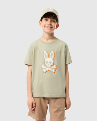 A smiling young boy wearing a light green **Psycho Bunny KIDS KAYDEN GRAPHIC TEE - B0U676C200** with a cartoon bunny skull and crossbones design. He is also sporting a matching light green baseball cap and beige shorts. The background is plain white.