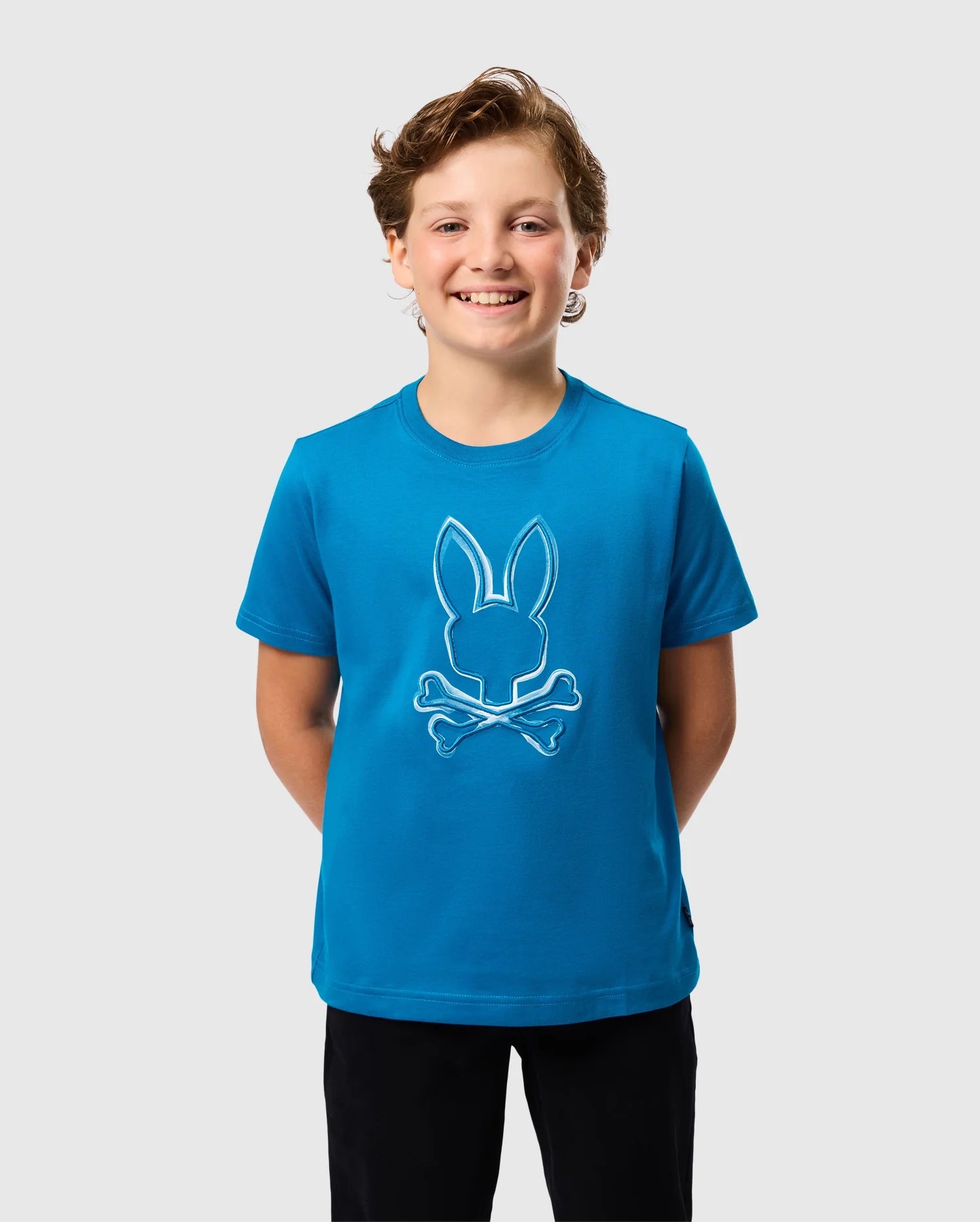 A smiling child wears the KIDS VINCENT GRAPHIC TEE - B0U713D200 from Psycho Bunny, featuring a bunny skull and crossbones design, against a plain light gray backdrop.