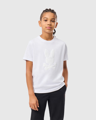 A young person with braided hair wears a white Kids Kingston Graphic Tee - B0U743D200 by Psycho Bunny, made from a soft Pima cotton blend. The T-shirt features an embossed center bunny graphic of a skull and crossbones. They are standing against a plain, light gray background with one hand in their pocket, looking at the camera with a neutral expression.