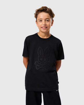 A young child with light skin and brown hair is wearing a Psycho Bunny KIDS MALTON GRAPHIC TEE (B0U795D200) in black, featuring a bunny skull and crossbones. The child stands against a plain white background, smiling with hands tucked in front pockets.