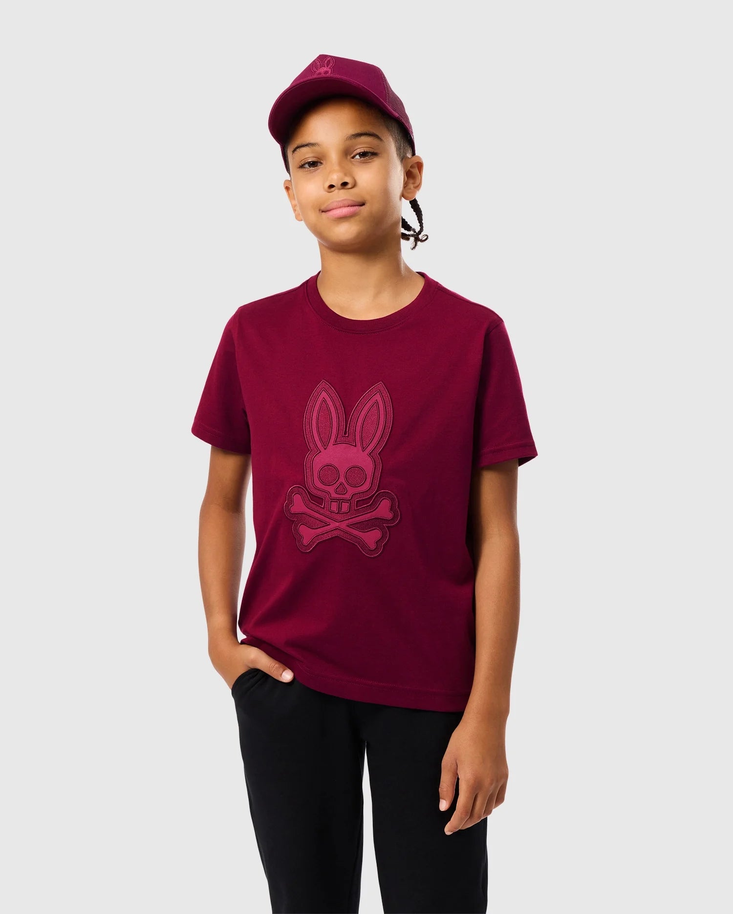 A young person stands confidently against a plain backdrop wearing a maroon Psycho Bunny KIDS MALTON GRAPHIC TEE - B0U795D200, featuring a design of a bunny skull and crossbones. With one hand in their pocket and a neutral expression, they showcase the comfortable kids' shirt effortlessly.