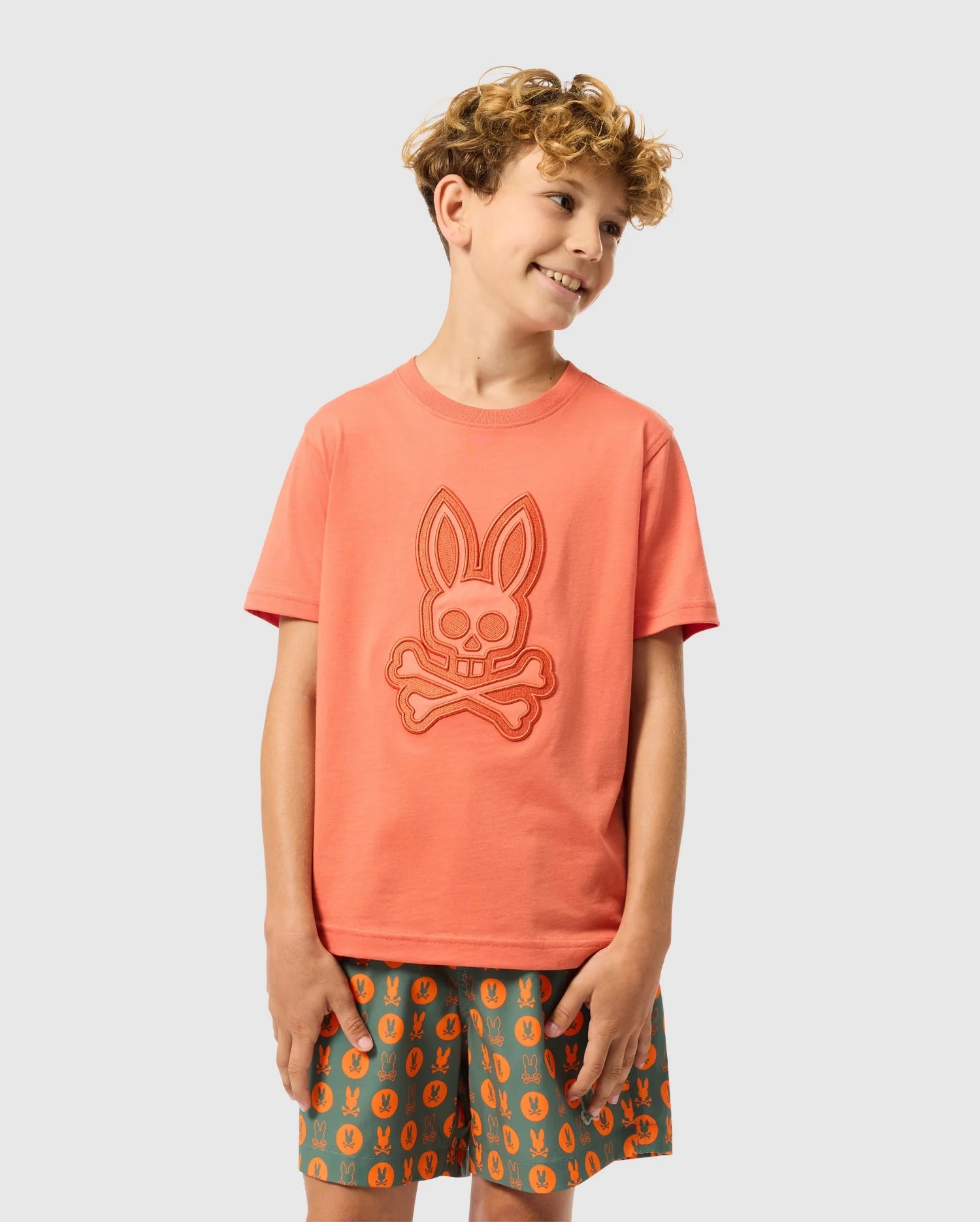 A young boy with curly hair is smiling and looking to the side. He is wearing a Psycho Bunny KIDS MALTON GRAPHIC TEE (product code: B0U795D200), a coral-colored Pima cotton shirt featuring a bunny skull and crossbones design on the front. He pairs it with green shorts showcasing a matching pattern. The background is plain light grey.