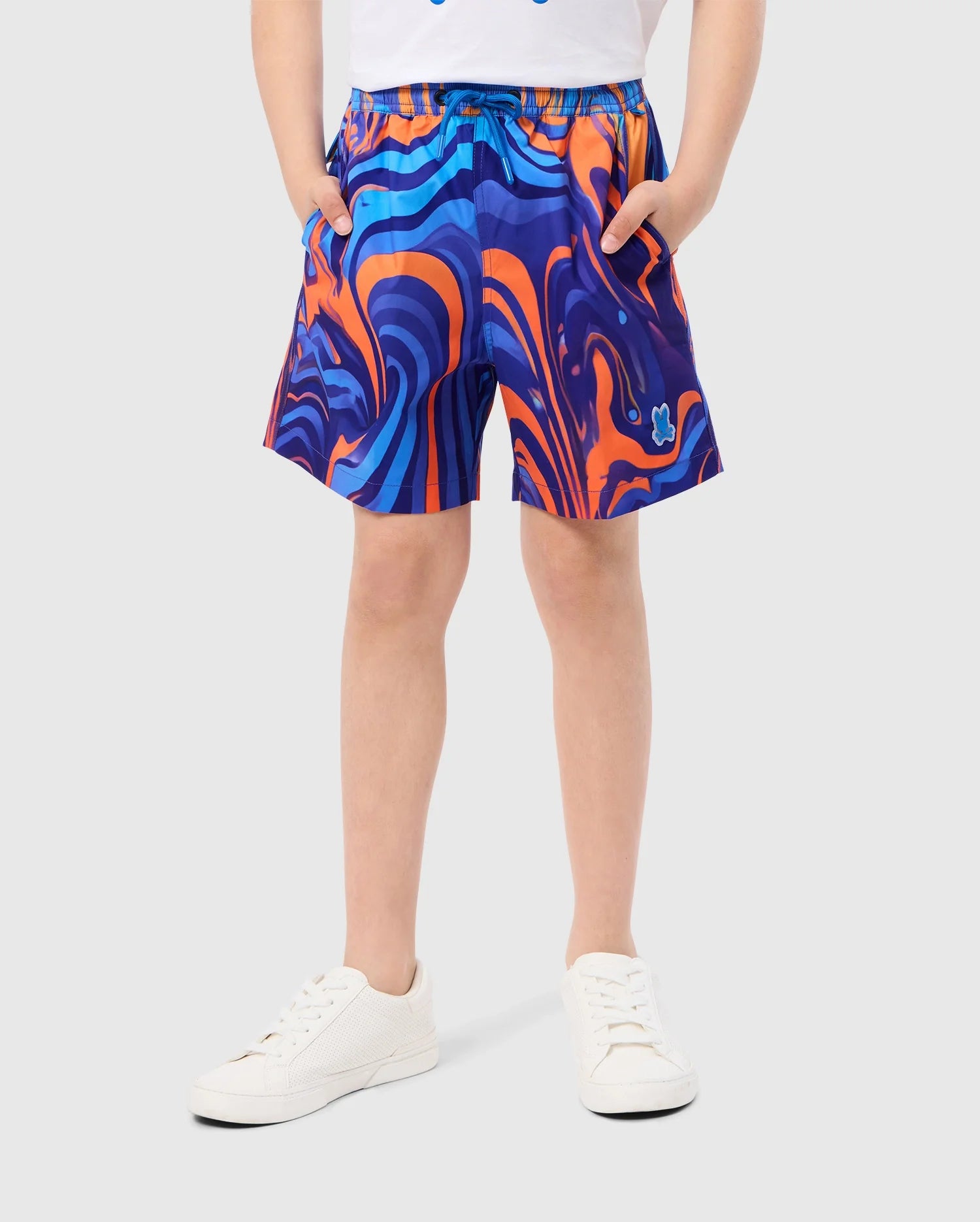 A person wearing KIDS NEVADA ALL OVER PRINT SWIM TRUNK by Psycho Bunny, which features a vibrant swirling blue and orange pattern, stands against a plain background in white sneakers. With their hands tucked into the pockets of the quick-dry fabric shorts, they exude a cool, casual vibe.