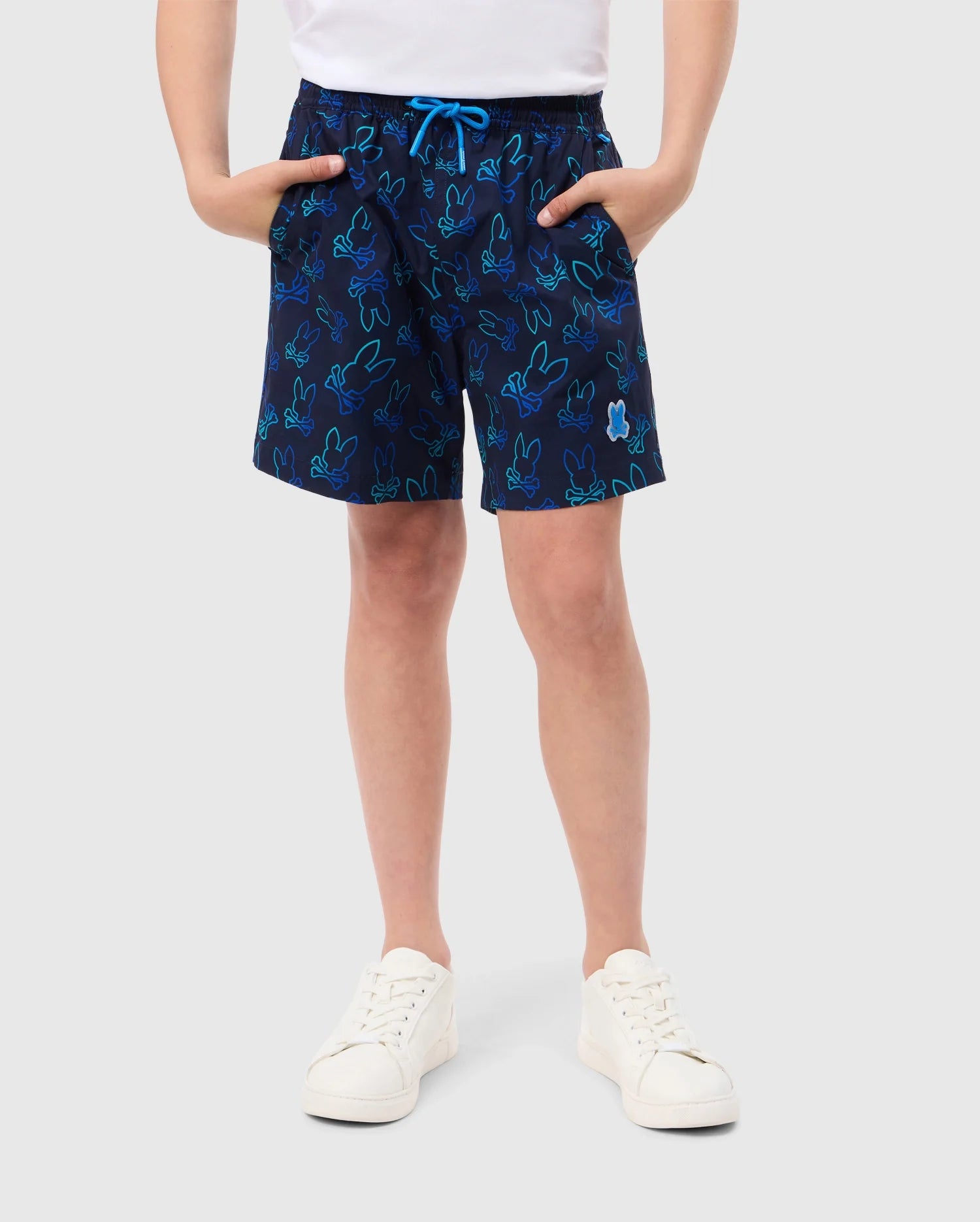 A person is shown from the shoulders down, wearing Psycho Bunny's KIDS SHELDON ALL OVER PRINT SWIM TRUNK - B0W588C200 in navy blue with a bunny pattern, a white drawstring, and a white patch. The person is also wearing a white t-shirt and white sneakers. Their hands are tucked into the pockets of the quick-dry fabric shorts.