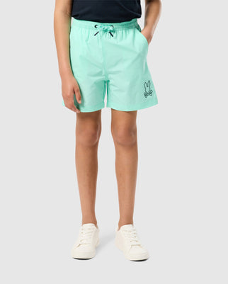 A child wearing light green, quick-dry KIDS PARKER HYDROCHROMIC SWIM TRUNK - B0W646C200 from Psycho Bunny with a small graphic of a bunny and crossbones on one leg stands confidently. The trunks have a drawstring waist. Paired with a dark shirt and white sneakers, the child is set against a plain, light grey background.