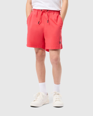 A person's lower half is visible, dressed in red KIDS PARKER HYDROCHROMIC SWIM TRUNKS by Psycho Bunny, featuring a small black bunny logo on the lower left side. They complement the look with white socks and white sneakers against a plain light gray background.