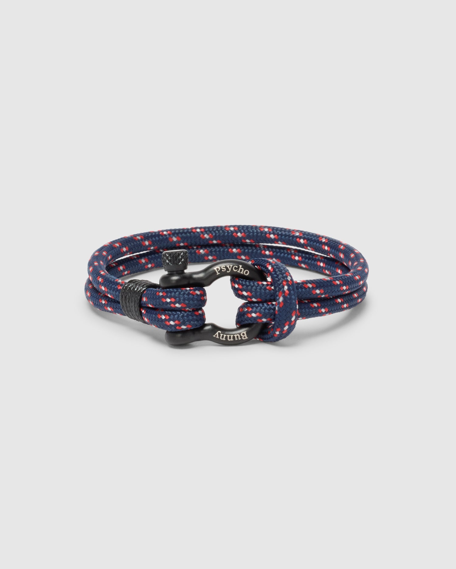 The Psycho Bunny MEN'S ROPE BRACELET - B6A368E200 is a stylish navy blue braided accessory with red and white speckles, featuring a modern lightweight design and an engraved metal clasp.