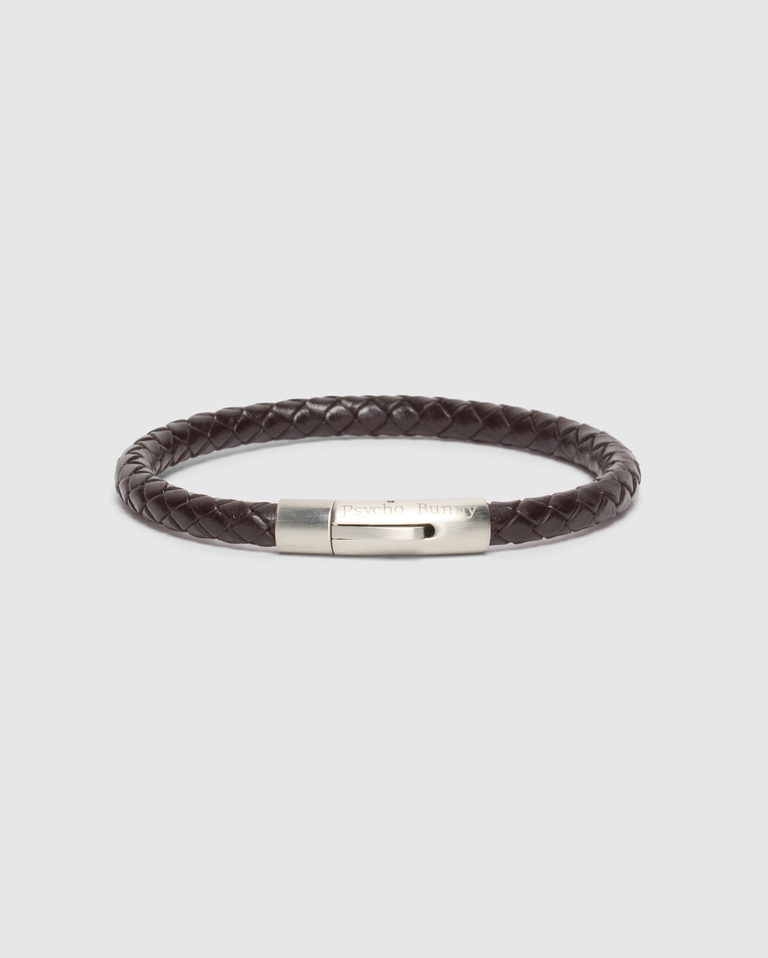 The Psycho Bunny Men's Braided Leather Bracelet - B6A371E200 features an elegantly eye-catching dark brown leather braid with a structured design and a silver engraved clasp, set against a plain light gray background.