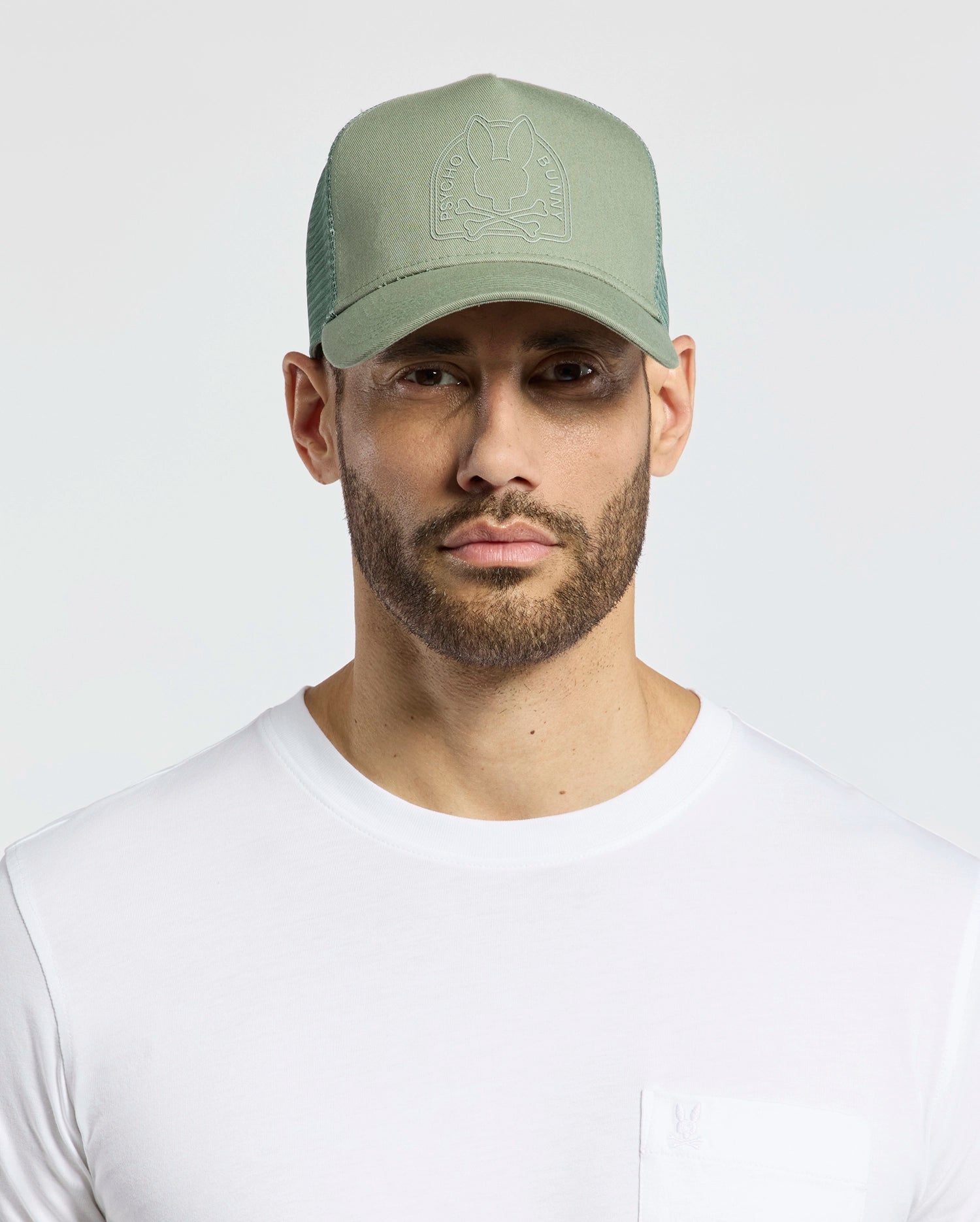 A bearded man wears a Psycho Bunny light green MENS MELBOURNE GRAPHIC TRUCKER HAT - B6A548F200 with an embossed logo and snap-back strap. His white t-shirt completes the look as he gazes into the camera, highlighting the hat's breathable design against a plain background.