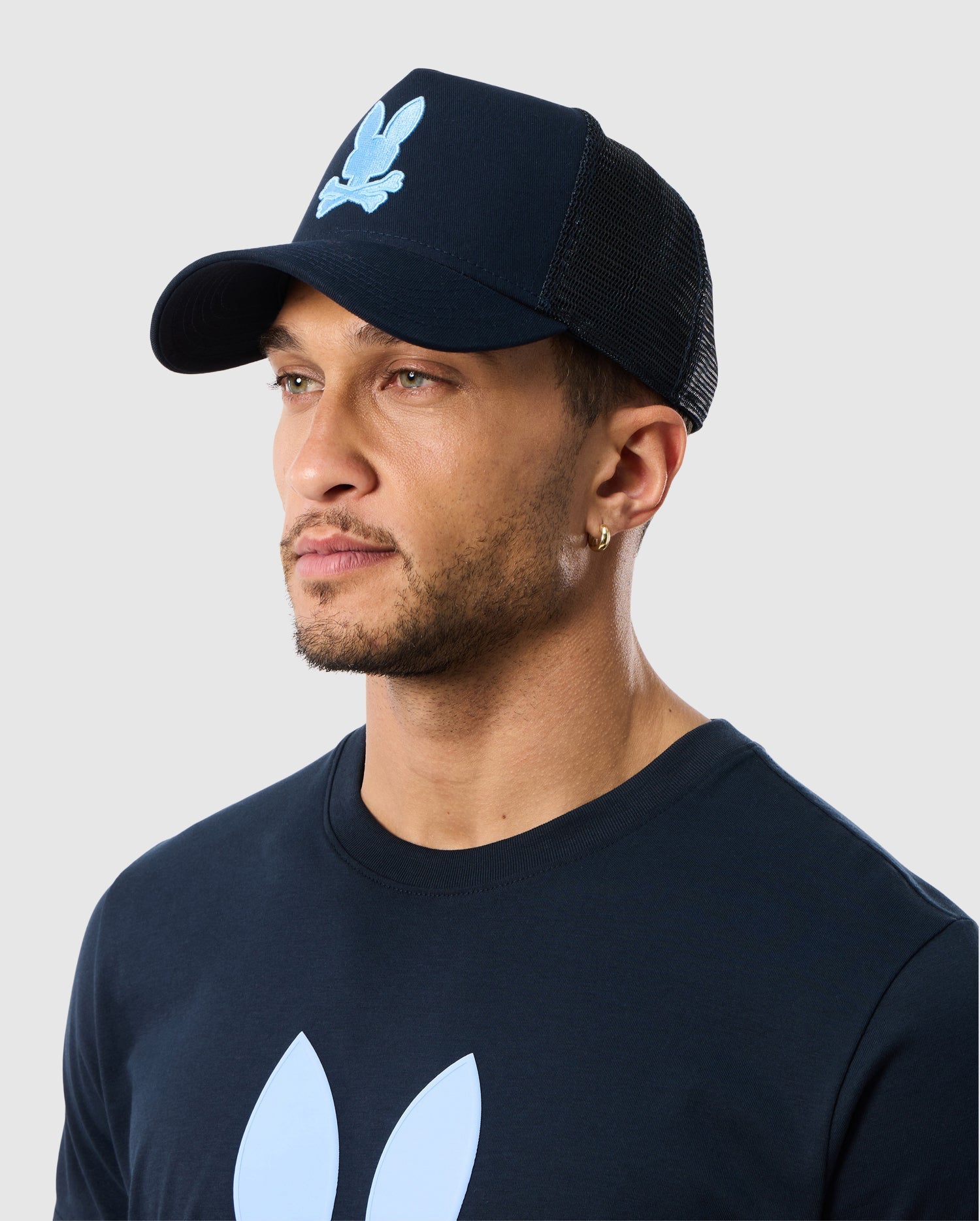 A person wearing a navy blue Psycho Bunny MENS HOUSTON TRUCKER HAT - B6A550C2HT with a light blue graphic on the front, matching a navy blue t-shirt with a similar graphic. The individual has short hair and a beard, and they're looking off to the side against a plain, light gray background.