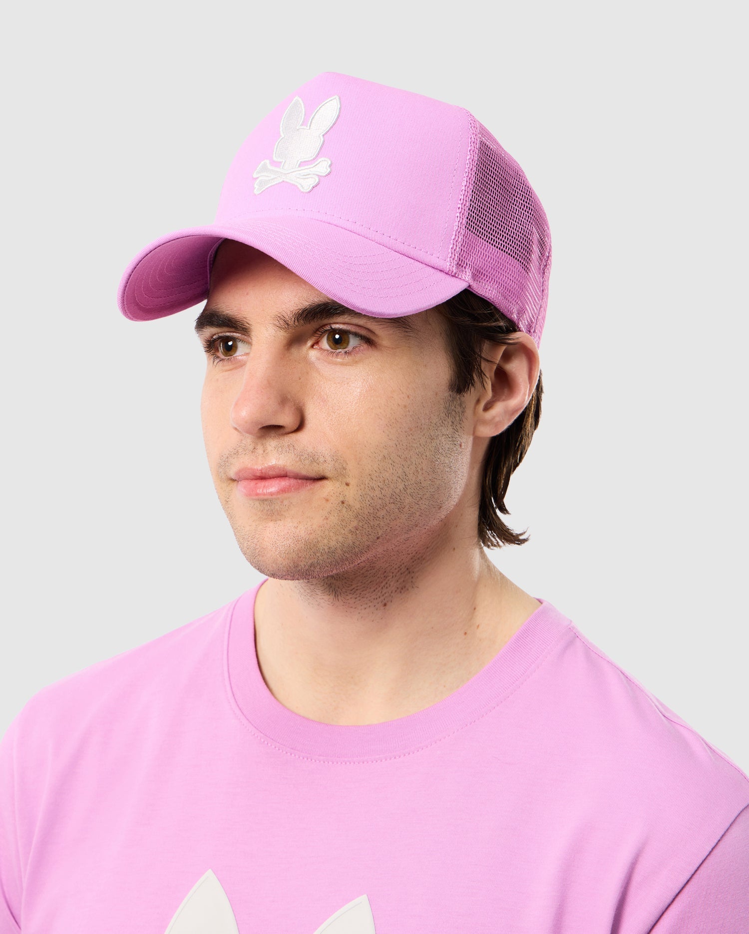 A person with short dark hair is wearing a pink Psycho Bunny MENS HOUSTON TRUCKER HAT - B6A550C2HT, featuring a white emblem of a bunny with crossed bones underneath. The matching pink t-shirt complements the look. The hat has snapback fastening and embroidered eyelet vents, set against a plain gray background.