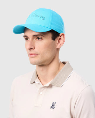 A person wearing a Psycho Bunny MENS LOUISE BASEBALL CAP - B6A551C200, made of durable cotton twill, and a beige polo shirt with a small bunny logo. The cap features an embroidered logo design that reads "bunny." The individual has a neutral expression and is posed against a plain gray background.