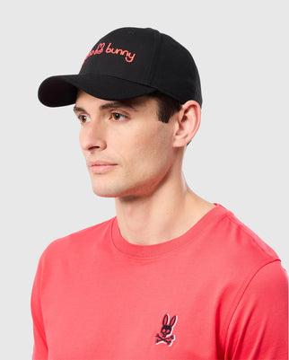 A man wearing the Psycho Bunny MENS LOUISE BASEBALL CAP - B6A551C200 in black, featuring an embroidered red bunny logo and a red T-shirt with a small bunny logo on the chest. The background is plain light grey.