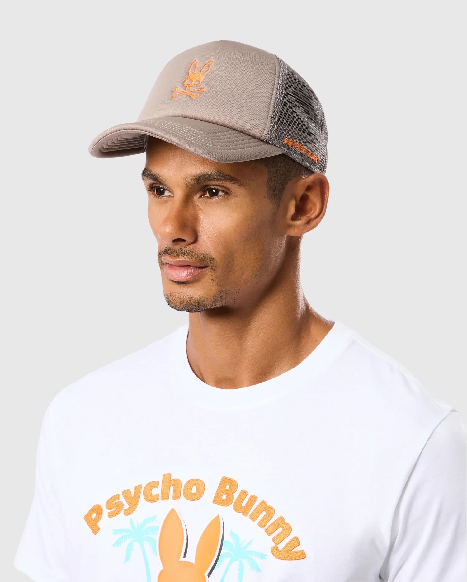 A person in a Psycho Bunny MENS PARIS TRUCKER CAP featuring ventilation eyelets and an orange bunny logo, with a matching T-shirt displaying 