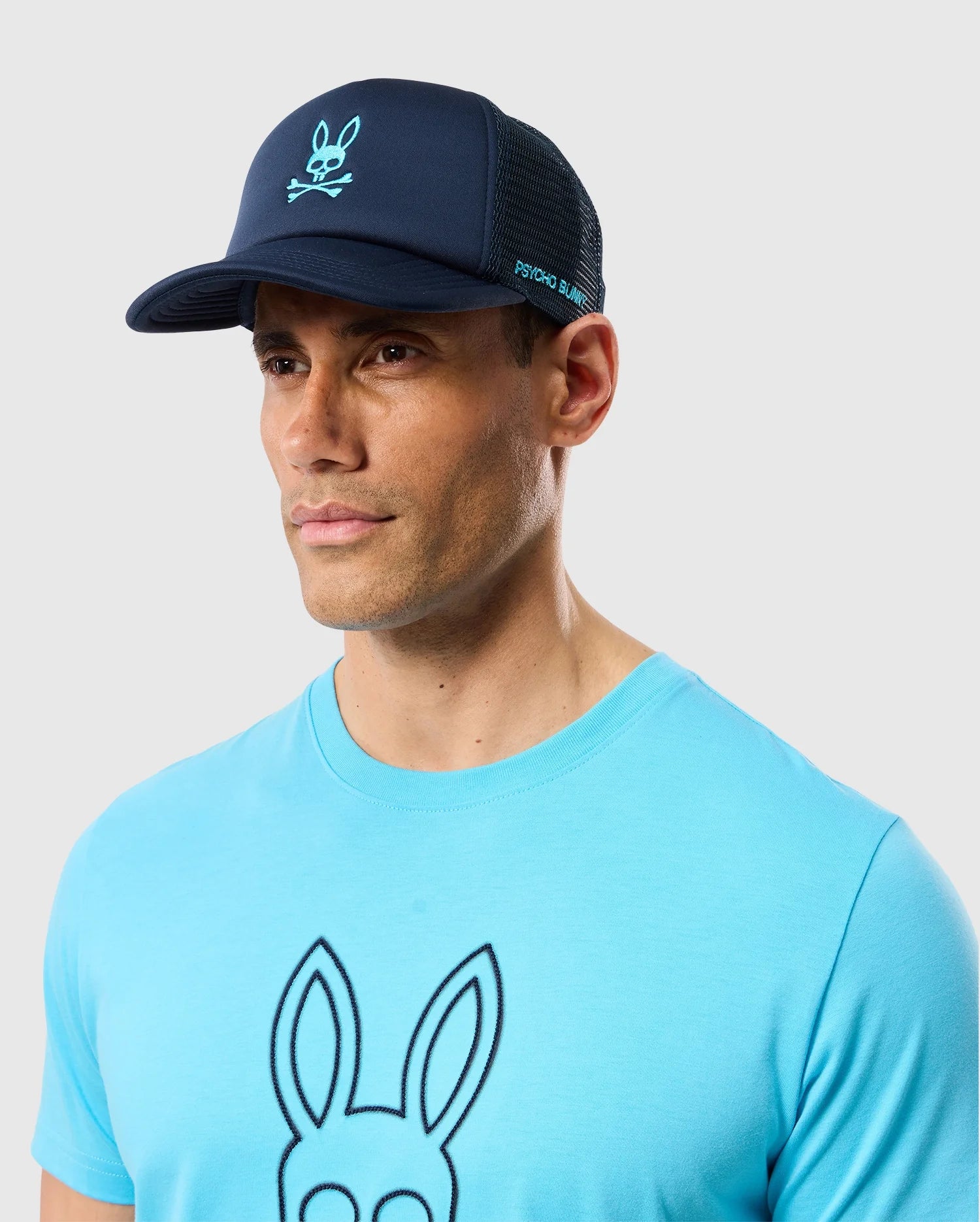 A man sporting a navy blue Psycho Bunny Paris Trucker Cap (B6A591C200), adorned with the distinctive Bunny logo and crossbones, perfectly complements his light blue t-shirt featuring the same iconic design on the chest. Set against a plain light gray background, this stylish ensemble promises all-day comfort.