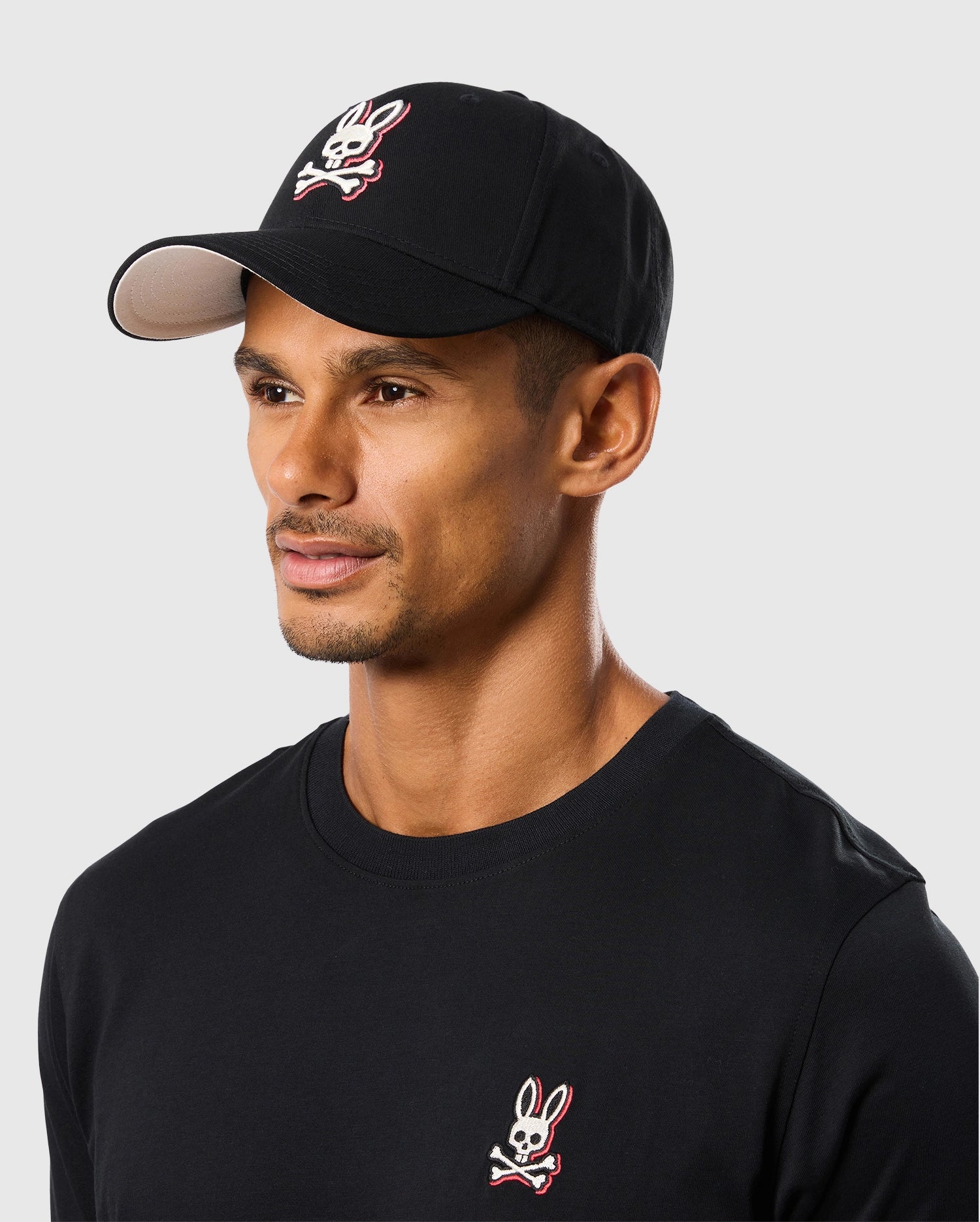 A man with a goatee is wearing the Psycho Bunny MENS KAYDEN BASEBALL CAP - B6A677C200 made from black cotton twill and a black long-sleeve shirt. Both the cap and shirt feature an embroidered white and red bunny skull and crossbones logo. He is looking off to the side against a plain light gray background.