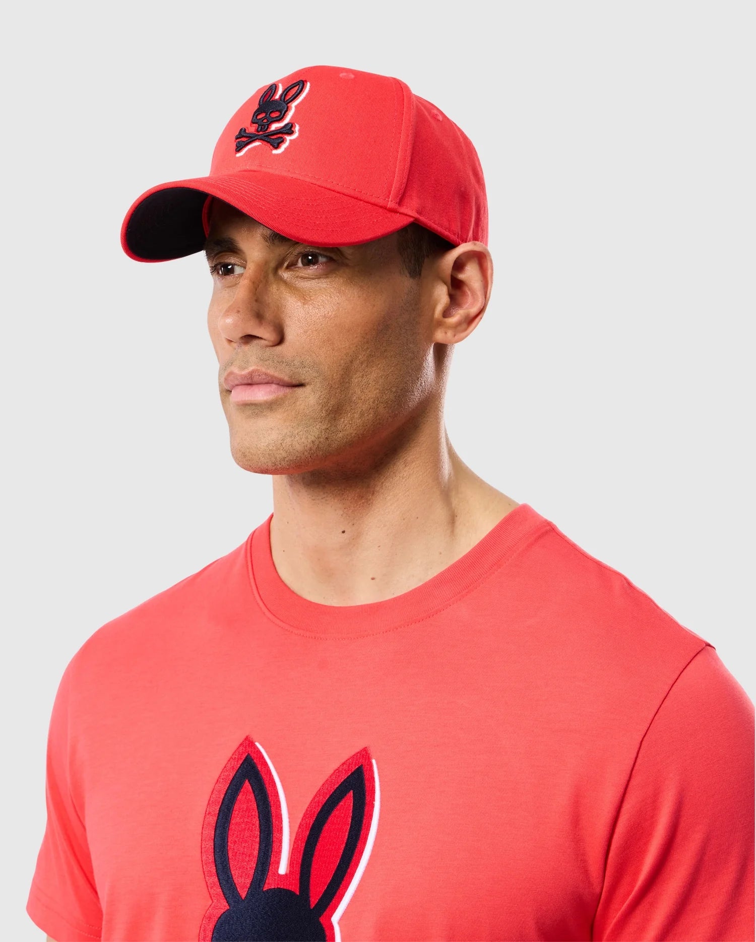 A man in a red Psycho Bunny Mens Kayden Baseball Cap (B6A677C200) and a matching T-shirt with the bunny logo stands against a light gray background.