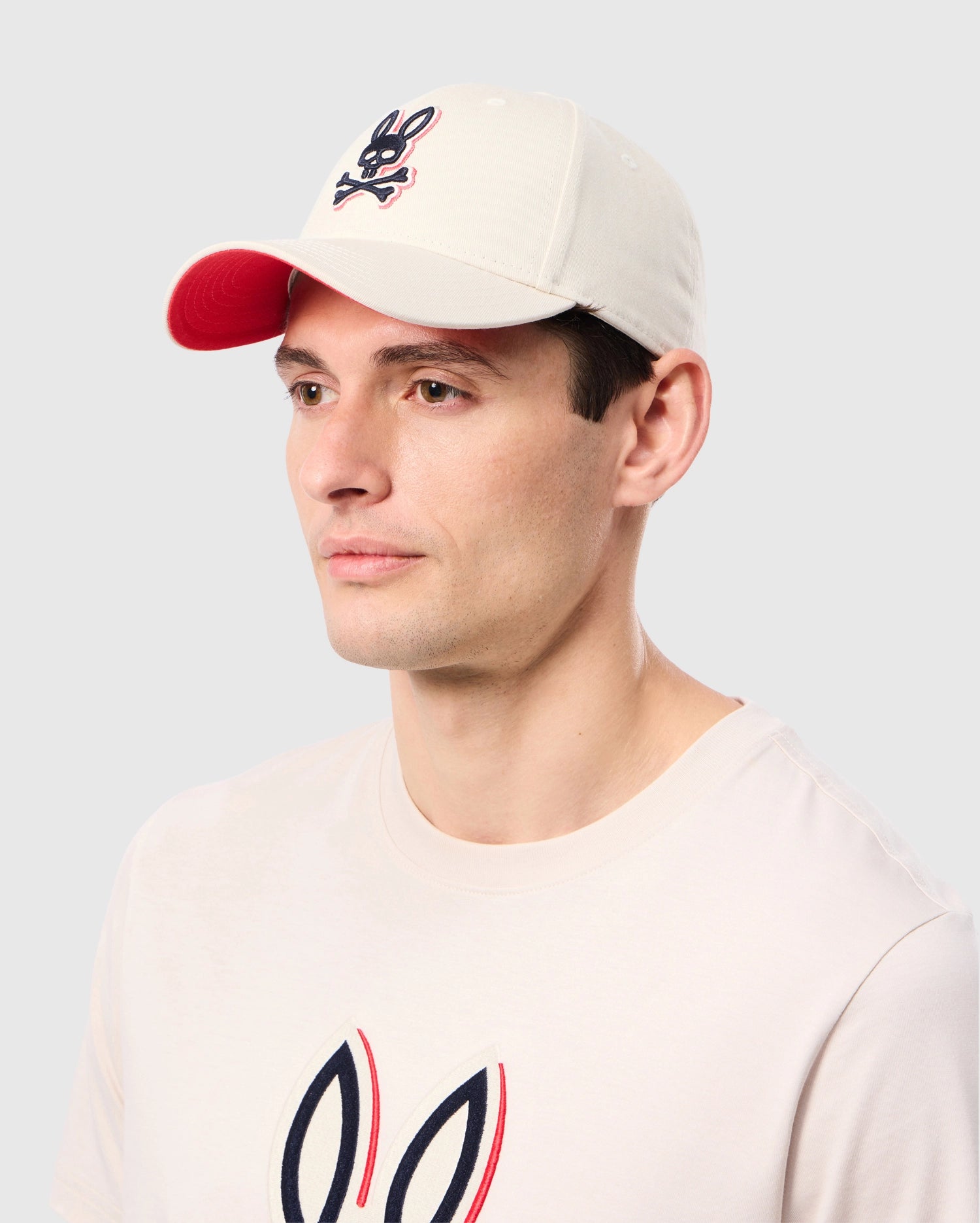 A man with light skin and short brown hair wears the MENS KAYDEN BASEBALL CAP - B6A677C200 by Psycho Bunny, a beige cap featuring a red brim and an embroidered black and red skull logo with bunny ears. He is also wearing a durable cotton twill beige T-shirt adorned with a similar black, red, and white bunny logo on the chest.