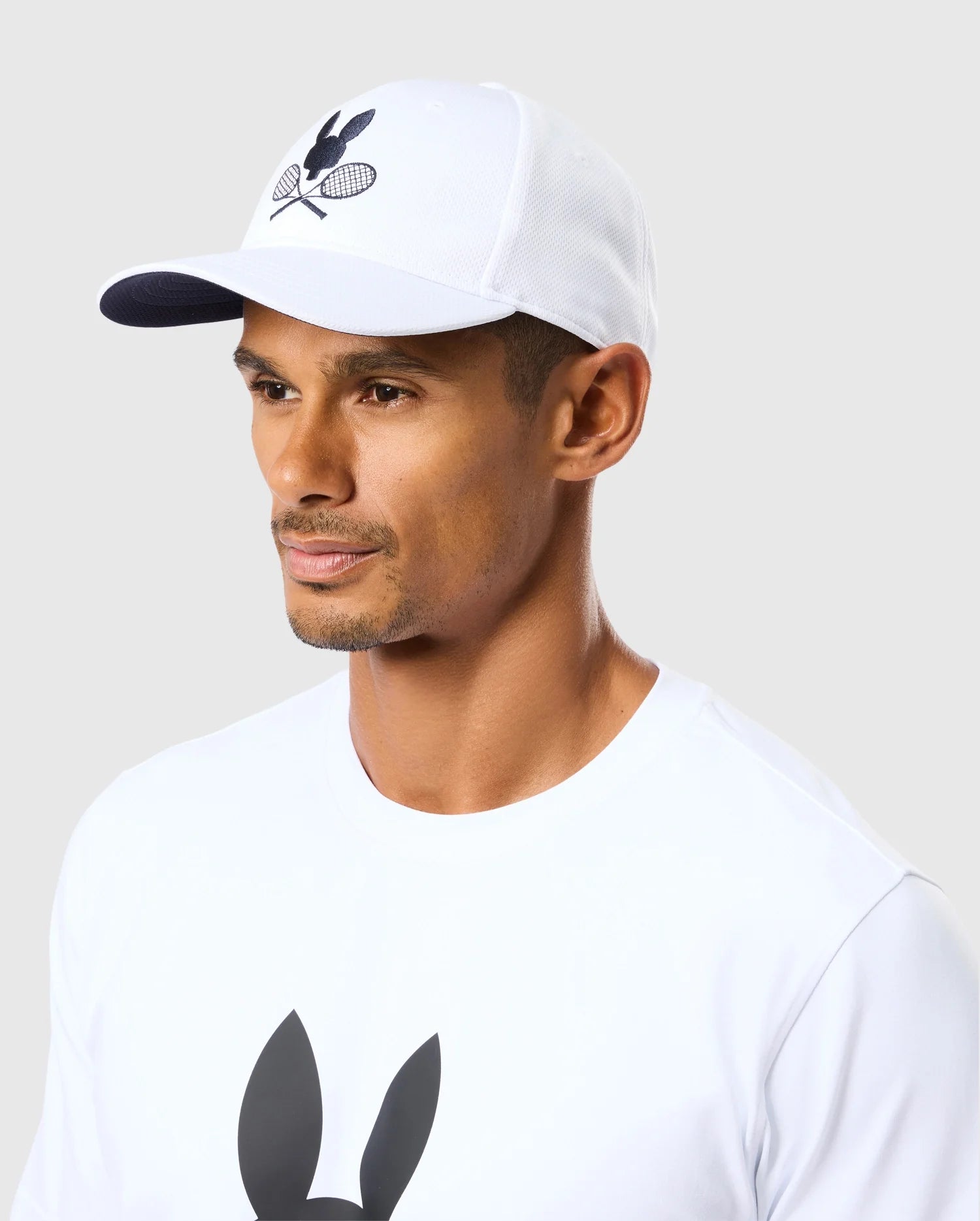 A person wearing the Psycho Bunny MENS COURTSIDE SPORT CAP - B6A685C200, featuring a tennis racquet and ball logo, paired with a white t-shirt showcasing a similar design. The light gray background beautifully accentuates the tennis-inspired outfit.