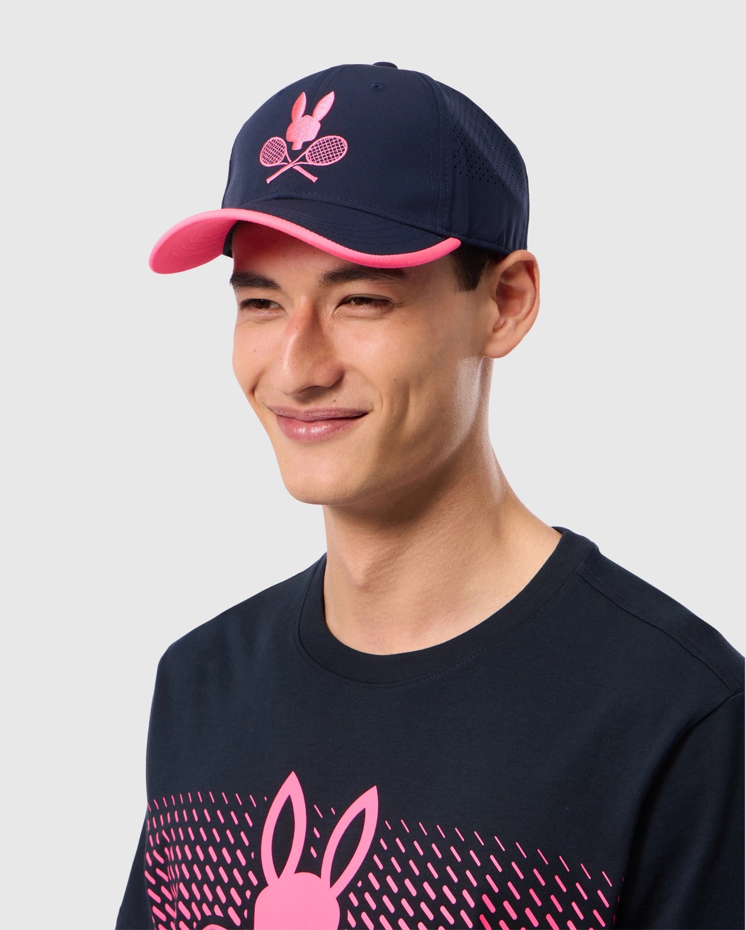 A young man smiles while wearing the Psycho Bunny MENS TORONTO TOURNAMENT CAP - B6A704C200, a navy blue baseball cap with a pink rim and an embroidered tennis bunny logo featuring crossed rackets. He is also sporting a matching navy blue shirt with a pink bunny graphic and geometric pattern.