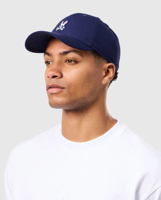 A person wearing a Psycho Bunny Men's Classic Baseball Cap in navy, featuring a white logo, and a white shirt stands against a light gray background.