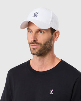 A bearded man wears Psycho Bunny's MENS CLASSIC BASEBALL CAP - B6A816A1HT, a classic white cap with adjustable fit, and a black T-shirt, both featuring the brand's small cartoon skull with rabbit ears logo against light gray.