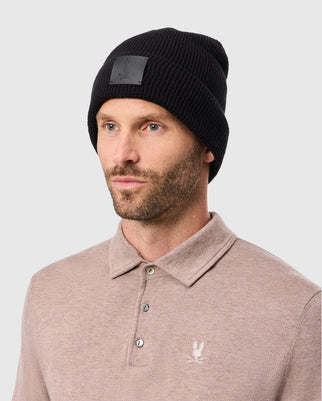 A man with a short beard is wearing a black Psycho Bunny MENS WAFFLE BEANIE (B6A873D200) and a light brown buttoned shirt with an embroidered logo on the chest. The background is plain light gray.