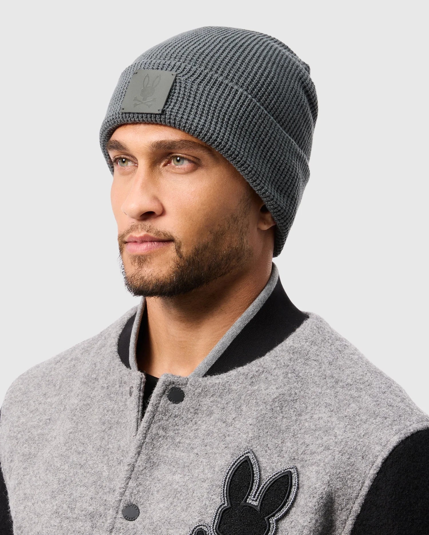 A man with light stubble is wearing a premium quality gray Psycho Bunny MENS WAFFLE BEANIE (B6A873D200) with a small patch and a gray varsity jacket featuring black trim and embroidered details. The background is plain light gray.