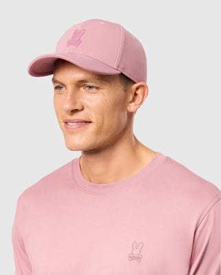 A man wearing a Psycho Bunny MENS JAMES PREMIUM BASEBALL CAP - B6A953D200 in pink polyester with an embroidered bunny design on the front, paired with a matching pink shirt featuring the same bunny logo. The background is light gray.