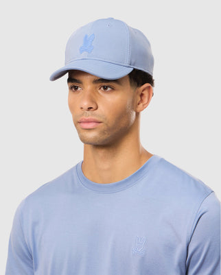 A person with short, curly hair is shown from the shoulders up, wearing a light blue Psycho Bunny Men's James Premium Baseball Cap (B6A953D200) made of knit twill fabric and a matching light blue T-shirt. Both the cap and T-shirt feature a small, tonal embroidered outline Bunny logo. The background is plain white.