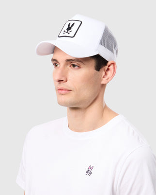 A person wearing a Psycho Bunny MENS BUNNY PATCH TRUCKER CAP with a breathable mesh back and black logo, paired with a white T-shirt featuring a small pink bunny logo. The background is plain gray.