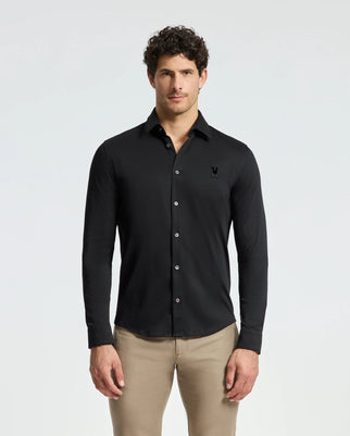 A man with curly hair is wearing the Psycho Bunny MENS INTERLOCK KNIT SHIRT - B6C188E200 in black and a pair of beige pants. Tailored from 100% Pima cotton, this long-sleeved button-up showcases a discreet embroidered logo on the chest. He stands against a plain white background.