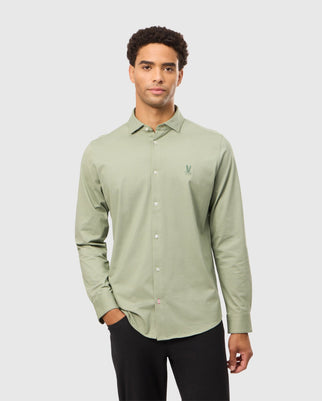 A man dressed in the Psycho Bunny MENS BRENDAN JERSEY SHIRT - B6C749D200, a light green button-up known for its high-quality craftsmanship and distinguished by a small embroidered emblem on the left chest, stands against a plain gray background with one hand in his black pants pocket and maintains a neutral expression.