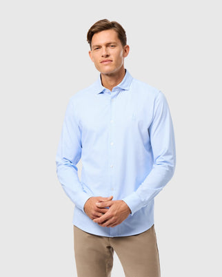 A man wearing Psycho Bunny's MENS BRENDAN JERSEY SHIRT in light blue, crafted from Pima cotton, paired with beige pants, stands against a plain white background. He has short brown hair and poses with his hands gently clasped in front of him, exuding a relaxed and confident expression.