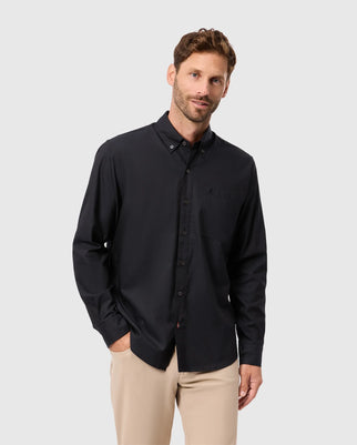 A man with short hair and facial hair is wearing a Psycho Bunny MENS JARED COTTON MODAL SHIRT - B6C774D200 with long sleeves and tan pants. He has one hand in his pocket and stands against a light gray background.