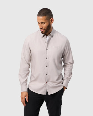 A man with short hair is wearing a light gray Psycho Bunny Jared cotton modal shirt and black pants. He stands against a plain background, looking down with one hand in his pocket.