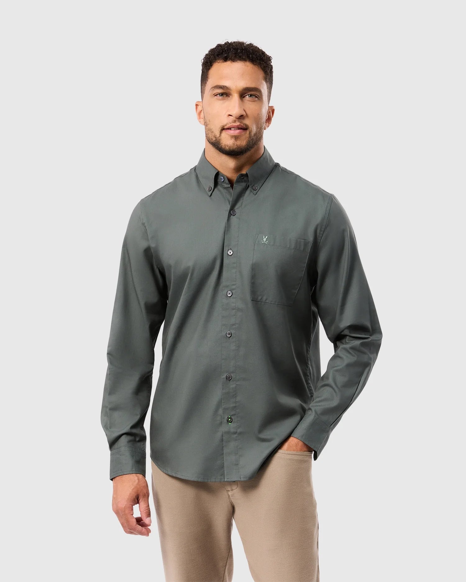 A man with short curly hair and a beard is wearing a dark green Psycho Bunny MENS JARED COTTON MODAL SHIRT - B6C774D200 and beige pants, standing against a plain gray background.