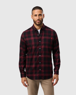 A man with short hair and a beard is wearing the MENS HAYES PLAID SHIRT - B6C842D200 in red and black by Psycho Bunny, made from 100% pima cotton. He complements it stylishly with beige pants, standing against a plain light gray background.