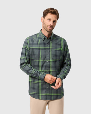 A man with short brown hair and a beard is sporting the trendy MENS HAYES PLAID SHIRT - B6C842D200 from Psycho Bunny, crafted from 100% pima cotton. He poses against a light gray background, gazing slightly to the side with his hands together, complementing his look with beige pants.