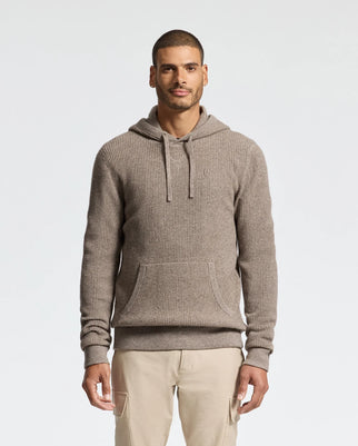 A man stands against a light background wearing the MENS LEVAN WAFFLE HOODED SWEATER - B6E246E200 by Psycho Bunny. The sweater features an embroidered chest bunny, ribbed cashmere-wool fabric, a front pocket, and is paired with beige pants, while his hands rest at his sides.