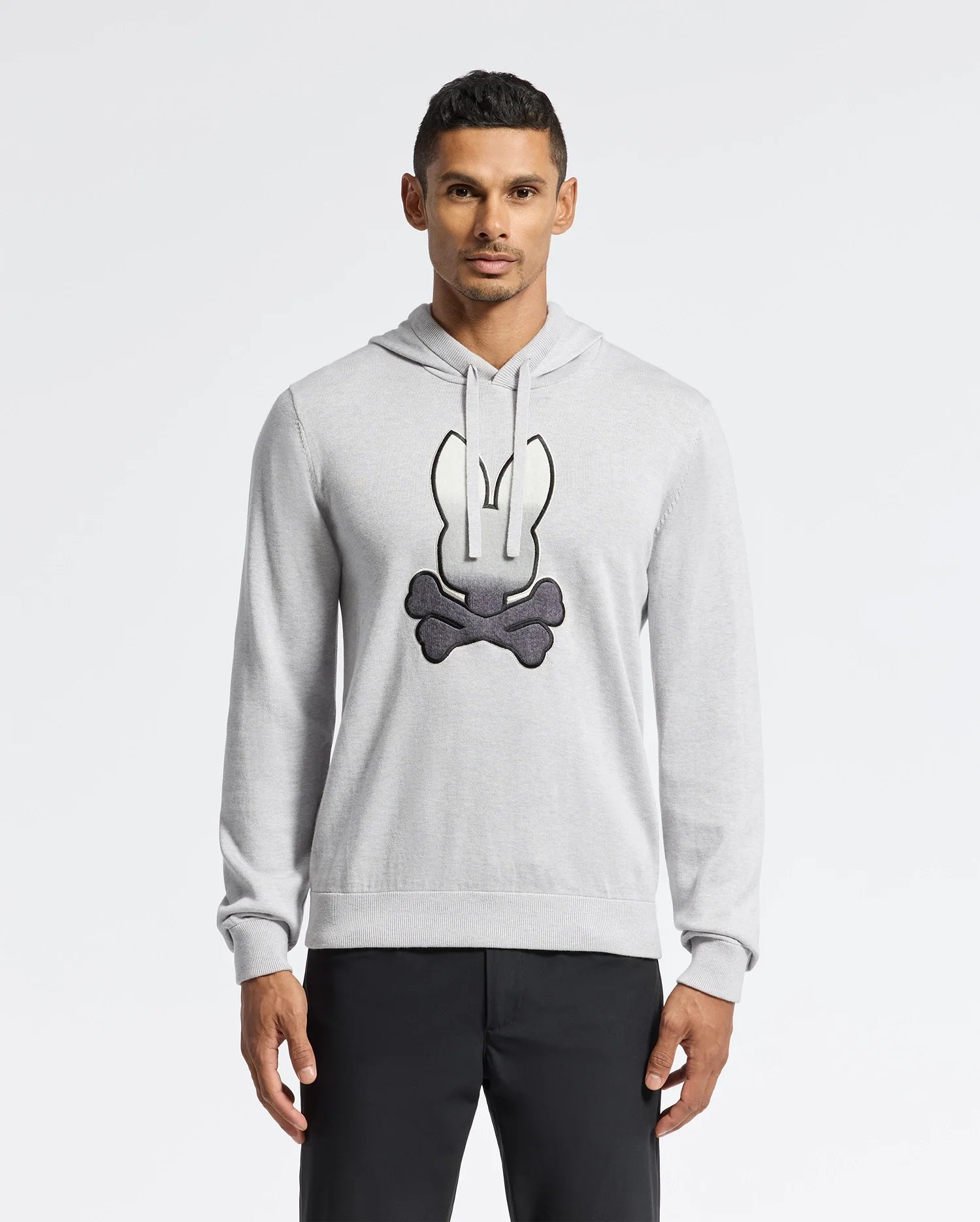 A man in a light gray Psycho Bunny MENS BULDER HOODED SWEATER - B6E261E200, showcasing a cartoon bunny and crossbones design, stands against a white background. Made from Supima Cotton, it pairs stylishly with his black pants for a comfortable look.