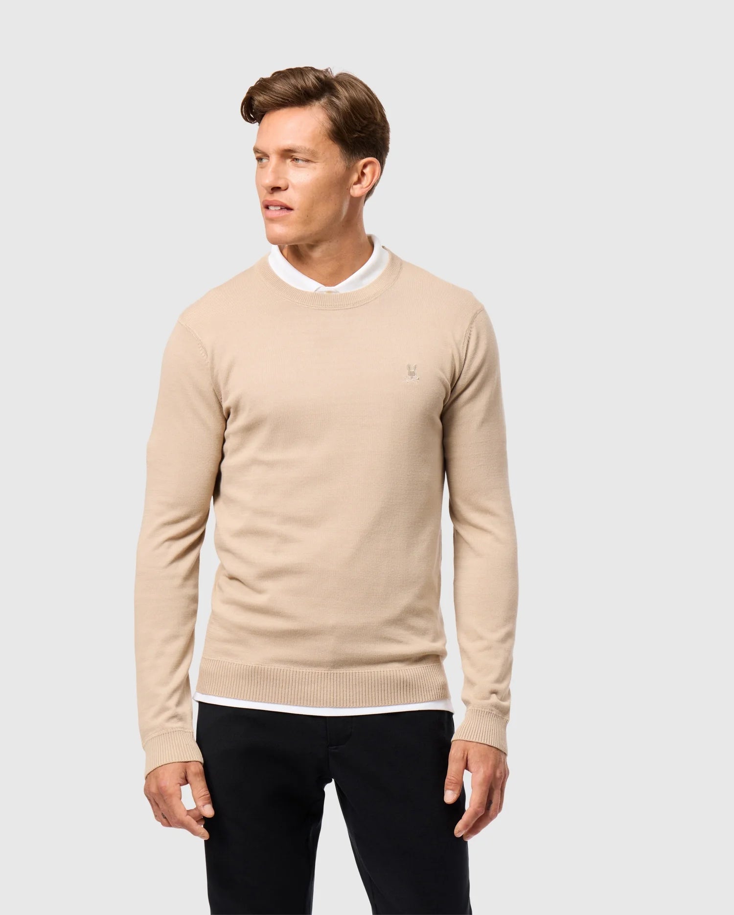 A person wearing the MENS BELMORE EXTRA FINE COTTON SWEATER from Psycho Bunny over a white collared shirt and black pants stands against a light gray background. The sweater subtly features an embroidered Bunny logo as they look to their left with a relaxed expression.