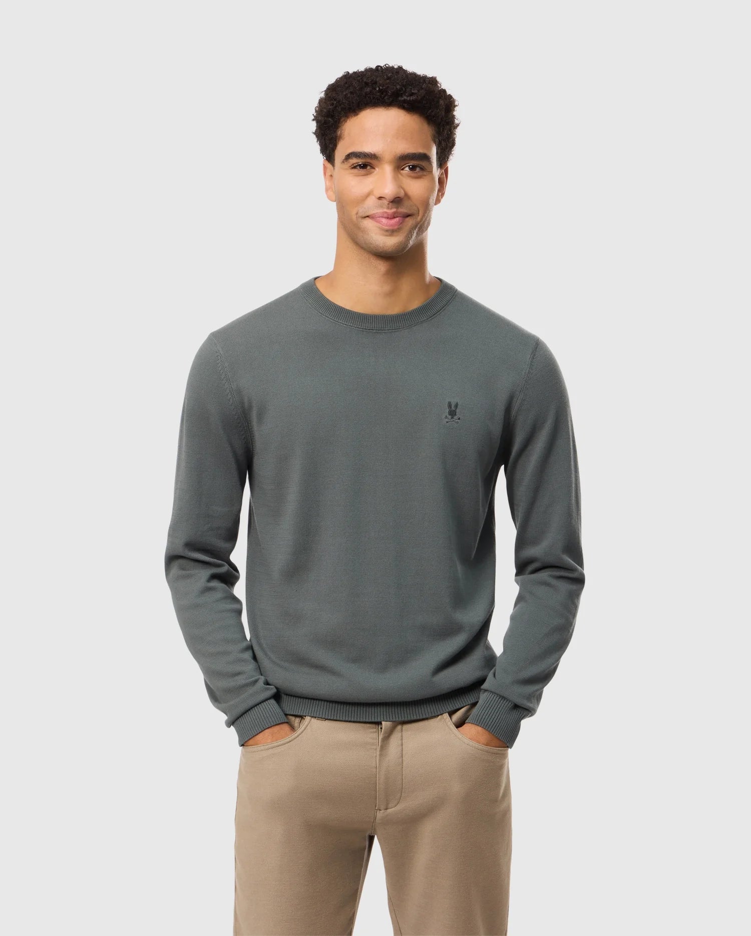 A man with short, curly hair stands against a plain white background. He is wearing the Psycho Bunny Men's Belmore Cotton Sweater - B6E832D200 in dark green and beige pants. The sweater features a small, embroidered logo on the left chest. With his hands in his pockets and a smile on his face, he exudes casual confidence.