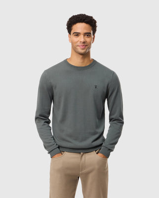 A man with short, curly hair stands against a plain white background. He is wearing the Psycho Bunny Men's Belmore Cotton Sweater - B6E832D200 in dark green and beige pants. The sweater features a small, embroidered logo on the left chest. With his hands in his pockets and a smile on his face, he exudes casual confidence.