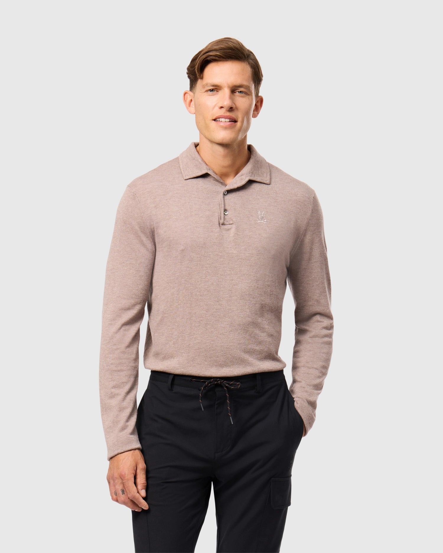 A person in a light brown Psycho Bunny MENS BRADSHAW POLO SWEATER (B6E835D200) paired with dark pants stands against a gray backdrop, smiling gently with hands by their sides, showcasing the elegance of luxury merino wool.