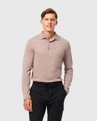 A person in a light brown Psycho Bunny MENS BRADSHAW POLO SWEATER (B6E835D200) paired with dark pants stands against a gray backdrop, smiling gently with hands by their sides, showcasing the elegance of luxury merino wool.